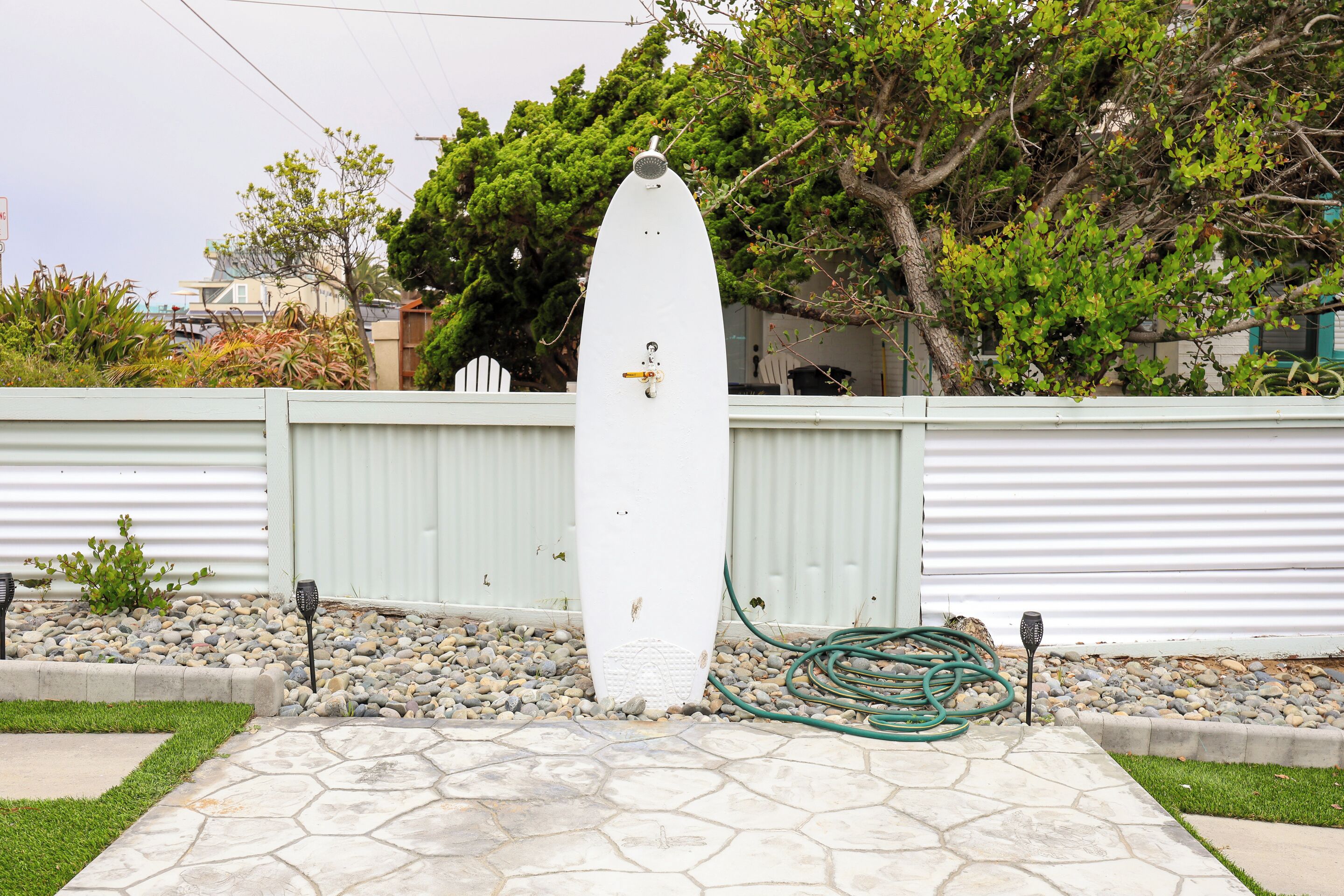 Une sculpture blanche en forme de planche de surf, située dans un espace jardin avec une terrasse en pierre, entourée d’une clôture blanche et de verdure.