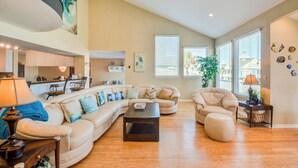 A spacious living room with a large L-shaped sofa, a round ottoman, and a coffee table. There is a kitchen area in the background, a dining table, and a large window with blinds. - Lei by the Sea, Beautiful 2nd Row Beach House with Elevator (Galveston)