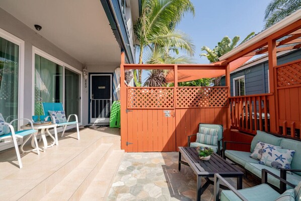 Une terrasse ensoleillée avec un espace salon, une clôture en bois et une porte portant l’inscription « Réservé au numéro 876 ».