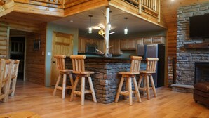 L’intérieur d’un chalet rustique, avec une cheminée en pierre, des tabourets de bar en bois, une cuisine dotée d’un plan de travail en pierre et un escalier en bois.