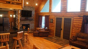 L’intérieur d’un chalet accueillant, avec une cheminée en pierre, des meubles en bois, un téléviseur et une grande fenêtre.