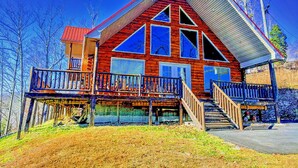 Une cabane en rondins dotée d’un toit à pignon bien visible, de grandes fenêtres et d’une terrasse en bois avec un escalier menant à un porche.