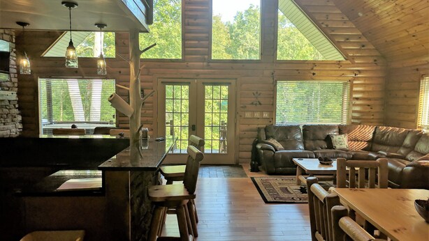 L’intérieur d’une cabane en rondins comprend un coin repas, un salon avec un canapé en cuir et de grandes fenêtres offrant une vue sur l’extérieur.