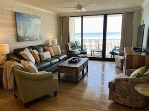 A spacious living room with a large window offering a view of the beach, a leather sofa, a coffee table, and a television.