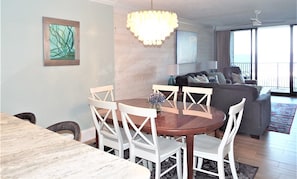 A dining area with a round table and white chairs, a chandelier, a painting on the wall, and a living area with a sofa and a TV.