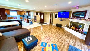 A spacious living room with a wooden floor, a TV mounted on the wall, a fireplace, and a kitchen in the background.
