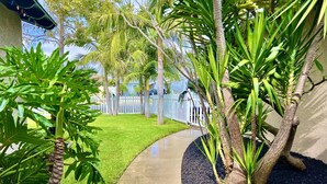 A pathway leading to a house with a white fence, surrounded by lush greenery and palm trees.