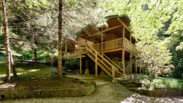 A wooden cabin with a wooden deck and stairs, surrounded by trees and a gravel path. - Private Creek, Hiking Trail System, Trout Fishing Onsite + Game Room, WiFi, Fire (Robbinsville)