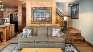 A cozy living room with a sofa, a coffee table with decorative items, a kitchen area with wooden cabinets, and a staircase. - Private Creek, Hiking Trail System, Trout Fishing Onsite + Game Room, WiFi, Fire (Robbinsville)