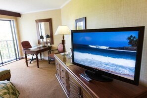A hotel room with a large flat-screen TV displaying a beach scene, a wooden desk with a lamp, a chair, and a bed with a green bedspread.