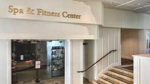 Eingangsbereich eines Spa- und Fitnessbereichs mit steinbelagtem Boden, einer Treppe mit Holzgeländer und einer Glastür zum Innenbereich.