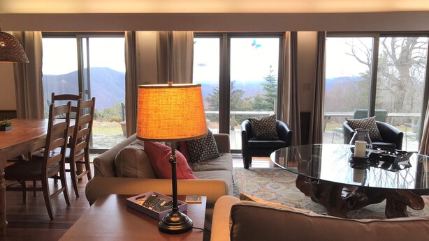 A cozy living room with a wooden table, a sofa, and a glass coffee table. There is a lamp on the table and a view of mountains through large windows. - Stunning Views, close to Wineries, Primland & Parkway - beautifully appointed (Meadows of Dan)