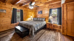 A wooden cabin bedroom with a bed, nightstands, a bench, a ceiling fan, and windows with curtains.
