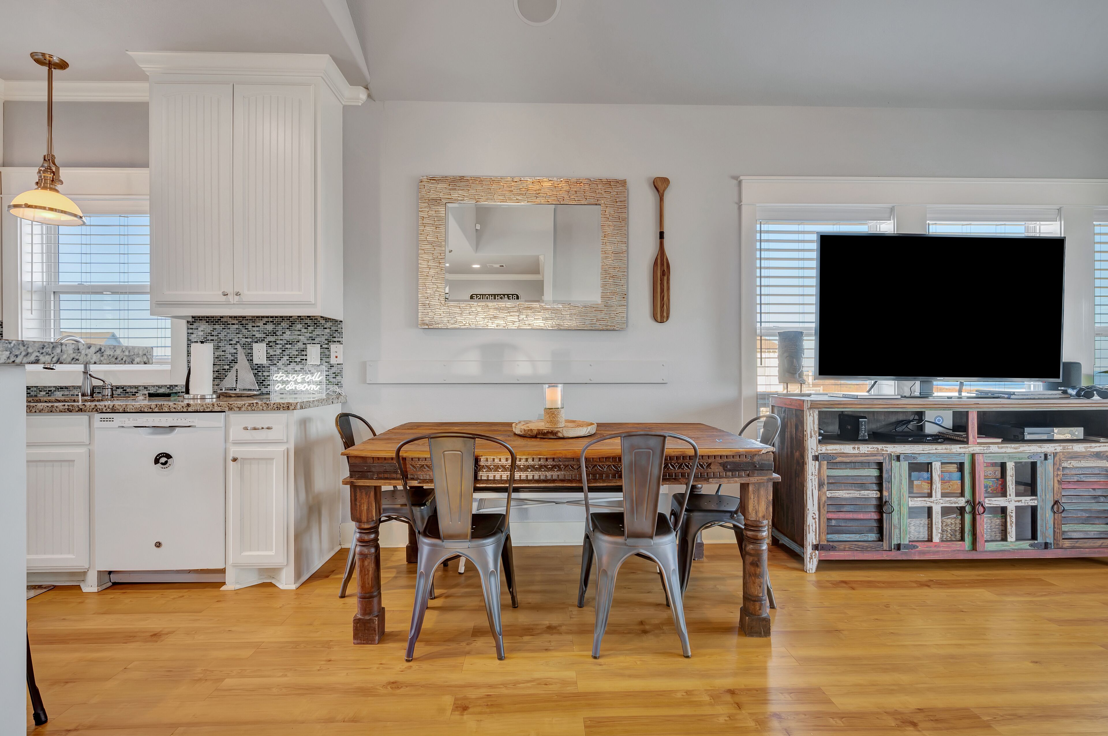 A modern kitchen with white cabinetry, a dining area with a wooden table and metal chairs, a large mirror, and a television on a wooden stand.
