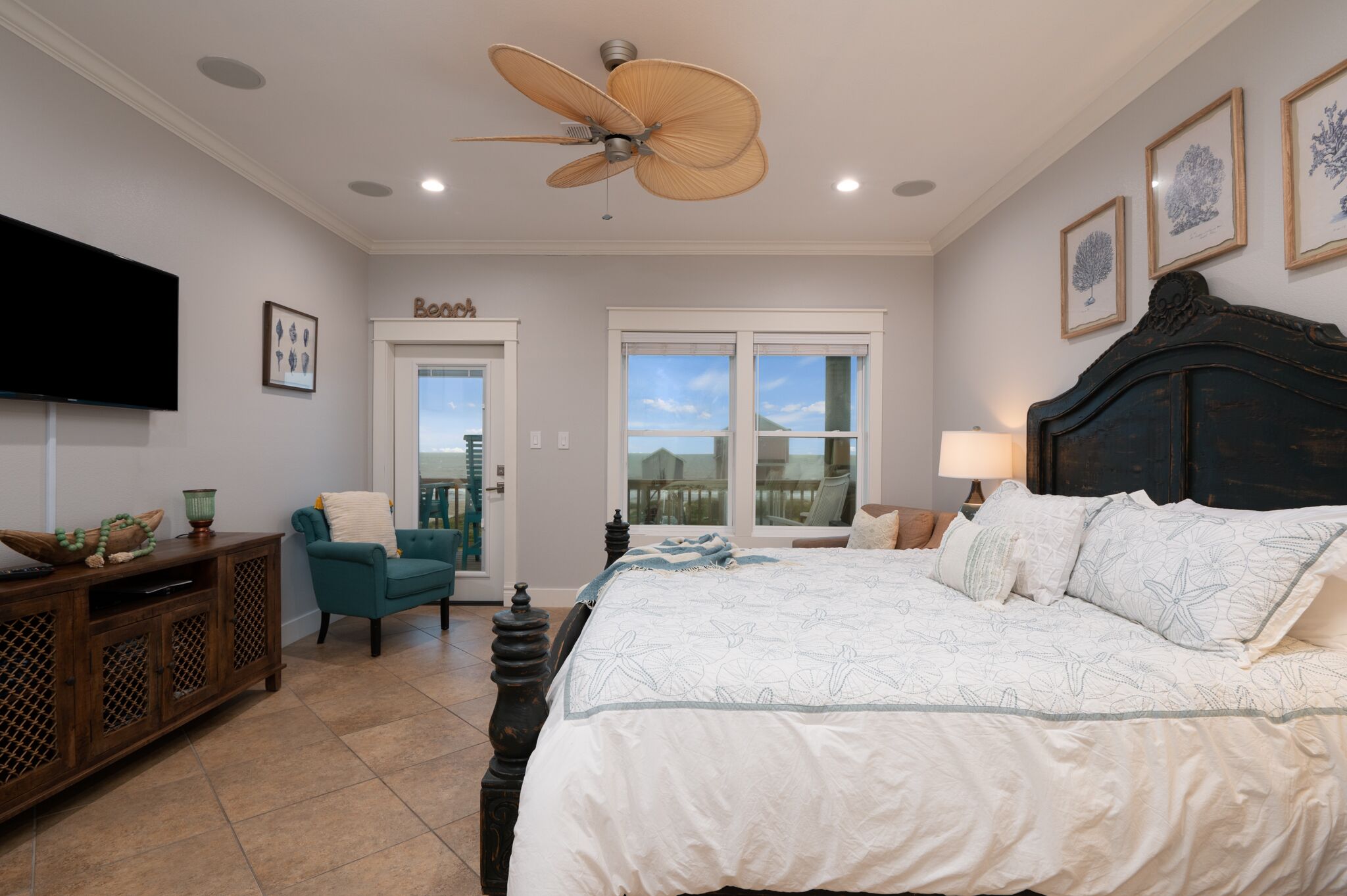 A bedroom with a large bed, a ceiling fan, a television, a desk, and a view of the outdoors through the windows.