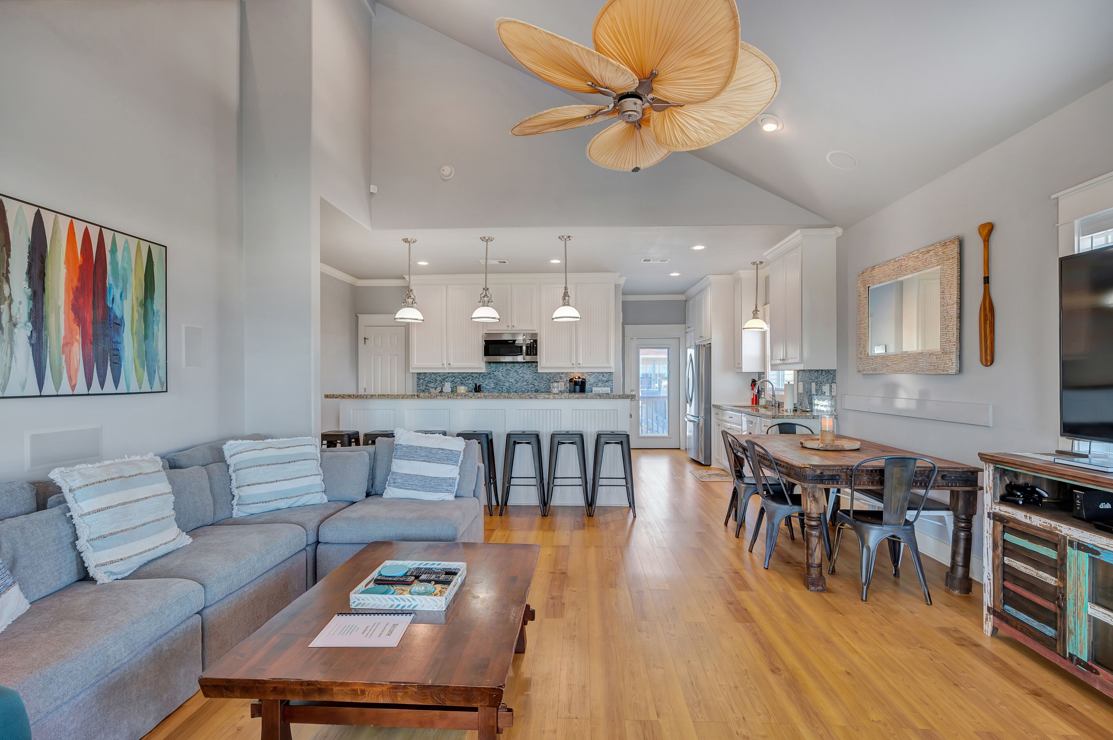 A spacious living area with a sectional sofa, a wooden coffee table, and a dining table with chairs. A ceiling fan and pendant lights are visible.