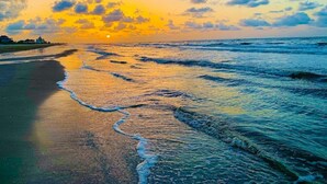 Une plage avec des vagues au coucher du soleil, un ciel dégagé et un bâtiment visible au loin.