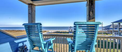 A wooden deck with two blue Adirondack chairs and a small table, overlooking a beach and a house.