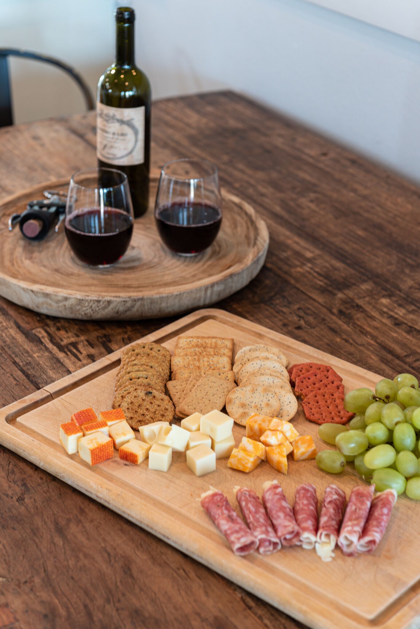 A wooden table with a charcuterie board featuring various cheeses, crackers, grapes, and cured meats. Two glasses of red wine and a bottle of wine are also visible.