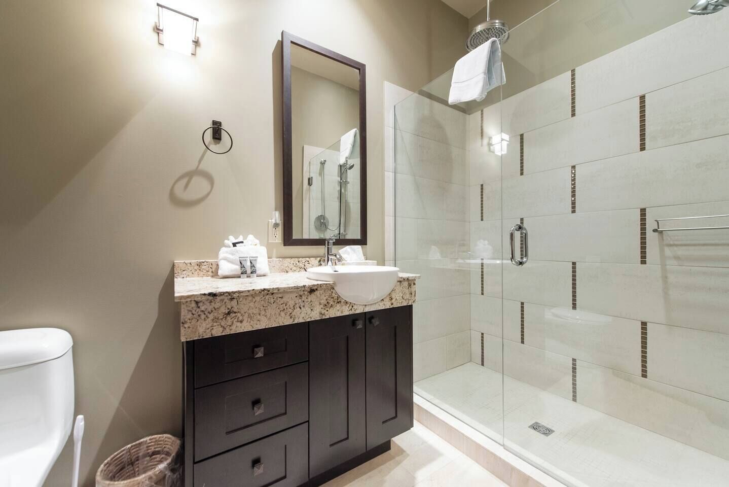 A modern bathroom with a granite countertop, a white sink, dark cabinets, a glass shower enclosure, and a large mirror.