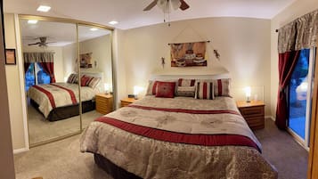 Une chambre à coucher avec un grand lit, deux tables de chevet, un ventilateur de plafond, un miroir et une fenêtre avec des rideaux.