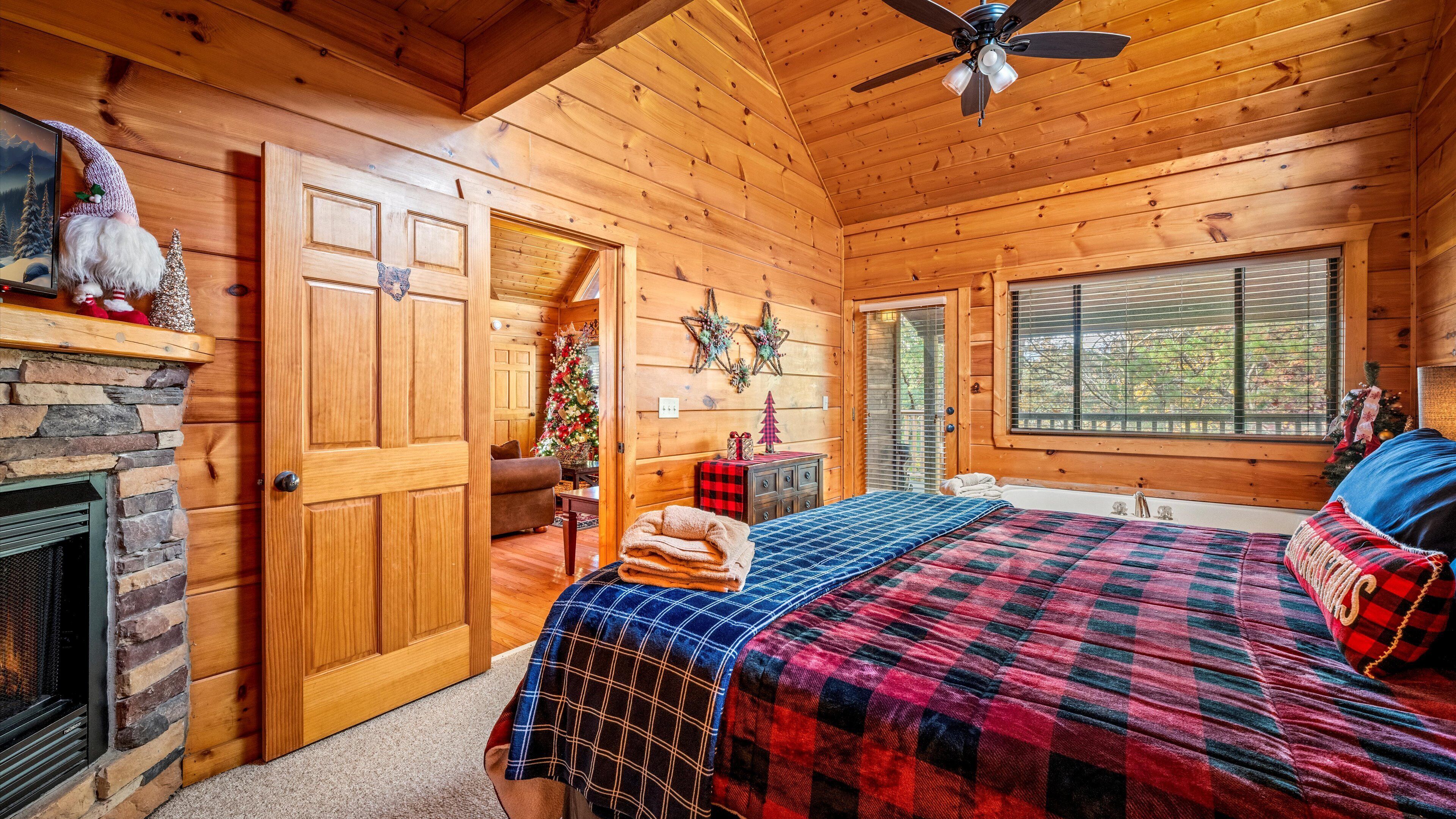 A cozy log cabin bedroom with a fireplace, a bed with plaid bedding, a ceiling fan, and a Christmas tree.