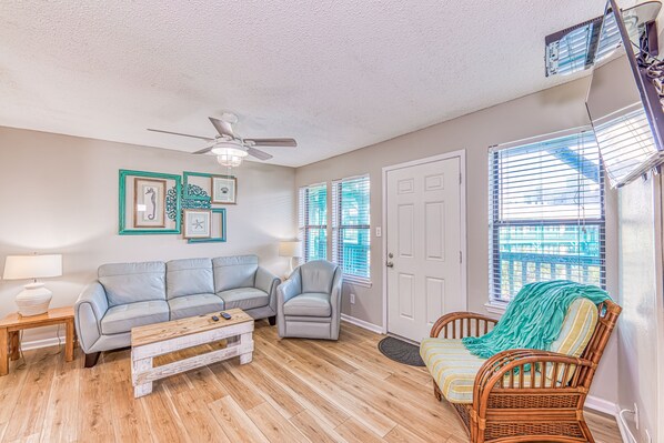 A living room with a light-colored sofa, a wooden coffee table, a wicker chair, and a ceiling fan. - Beach Area Condo, 2bed-2bath, Seabreeze 309 (Gulf Shores)
