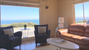 A room with a wooden ceiling, two armchairs, a sofa, a small table, and a lamp. Large windows offer a view of the sea and greenery.