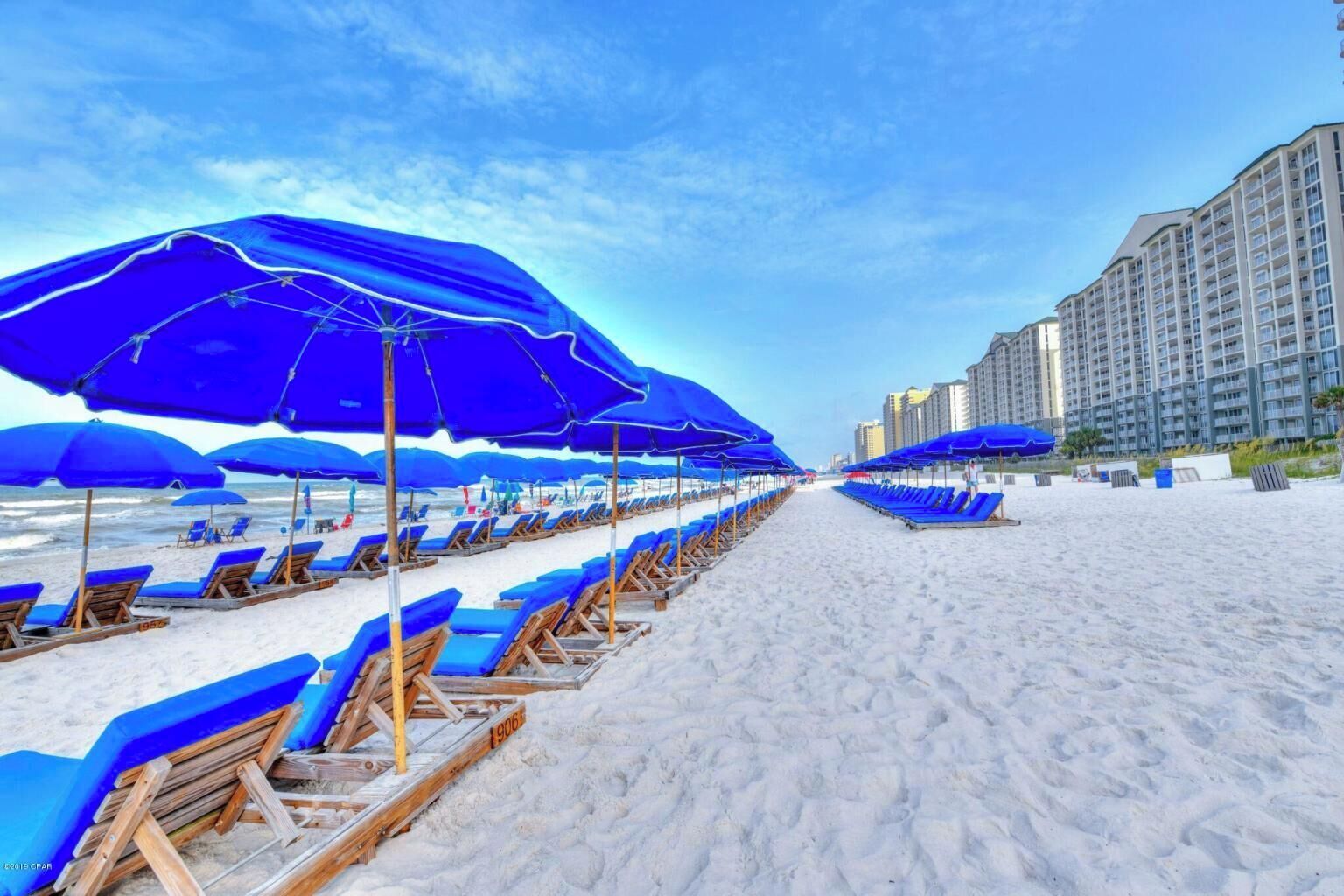 Una playa con hileras de sombrillas azules y sillas reclinables, un sendero arenoso y edificios altos al fondo.