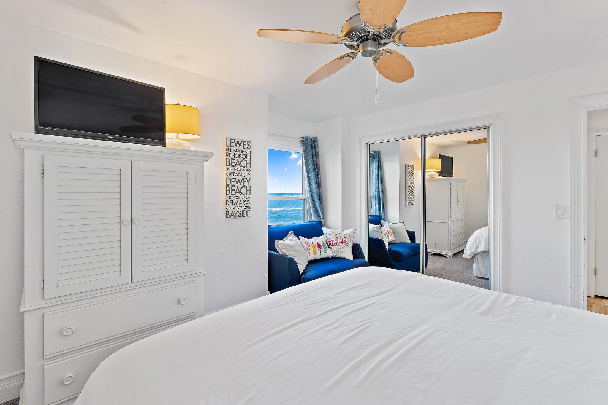 Une chambre avec un grand lit, un ventilateur de plafond, un téléviseur posé sur un meuble et une vue sur l’océan par une porte coulissante.