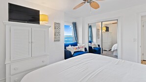Une chambre avec un grand lit, un ventilateur de plafond, un téléviseur posé sur un meuble et une vue sur l’océan par une porte coulissante.