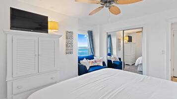 Une chambre avec un grand lit, un ventilateur de plafond, un téléviseur posé sur un meuble et une vue sur l’océan par une porte coulissante.