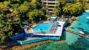 Vista aérea de un resort con una piscina grande rodeada de exuberante vegetación y un mar azul cristalino.