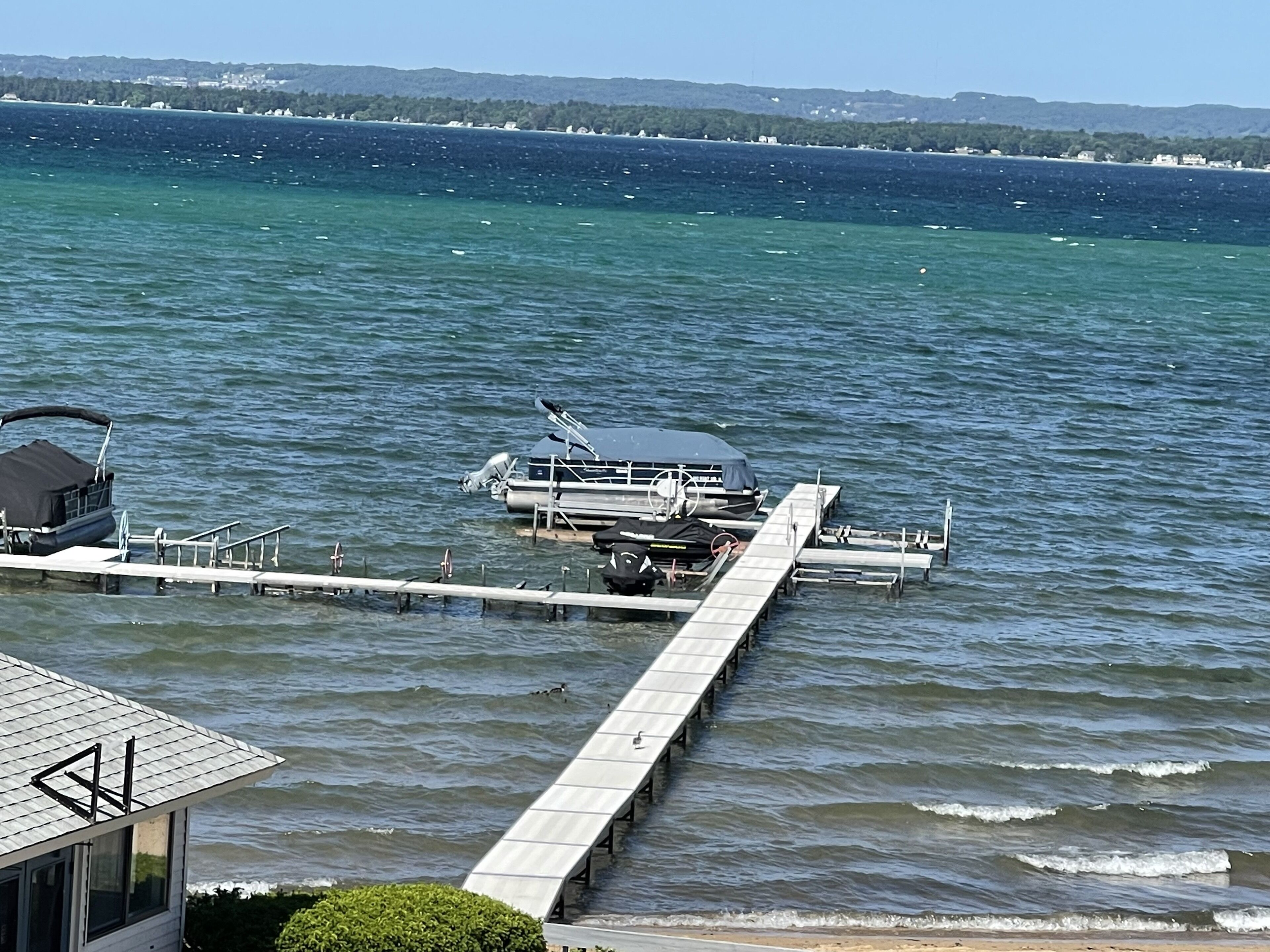 Un muelle se adentra en una masa de agua tranquila, con barcos amarrados, un cielo despejado y una costa distante con edificios.