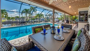 A poolside dining area with wicker furniture, a table set with plates, glasses, and a lantern.