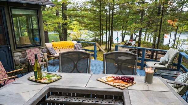 A deck with outdoor seating, a table with food and drinks, and a view of a lake surrounded by trees. - Quiet and Peaceful 3 Bedroom Family Cottage on Lake George, 12 Miles to TC (Traverse City)