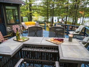 Eine Terrasse mit Sitzgelegenheiten im Freien, ein Tisch mit Speisen und Getränken und Blick auf einen von Bäumen umgebenen See.