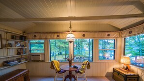 A cozy room with a wooden table, two chairs, a chandelier, a bookshelf, and large windows with a view of trees. - Quiet and Peaceful 3 Bedroom Family Cottage on Lake George, 12 Miles to TC (Traverse City)