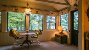 A cozy room with a wooden ceiling, a round table, two chairs, a lamp, and a view of trees through large windows. - Quiet and Peaceful 3 Bedroom Family Cottage on Lake George, 12 Miles to TC (Traverse City)