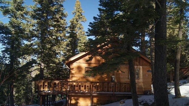 Una cabaña de madera con terraza, rodeada de altos pinos y nieve en el suelo.