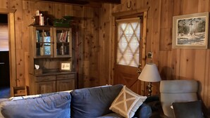 Un interior de cabaña acogedor con un sofá azul, una mesa de centro de madera, una estantería y una ventana con persianas.