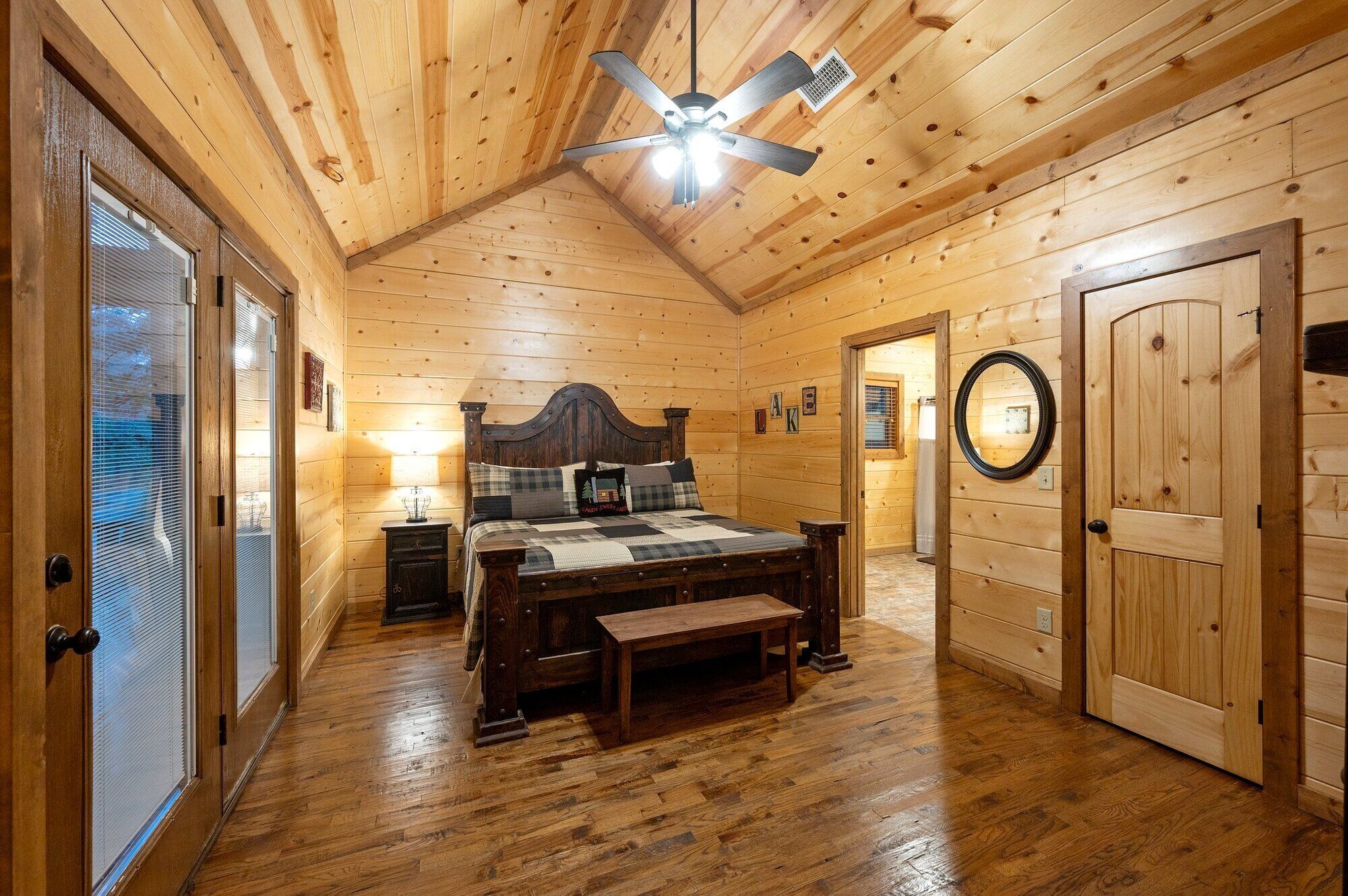 A wooden cabin bedroom with a bed, bedside tables, a ceiling fan, and a door.