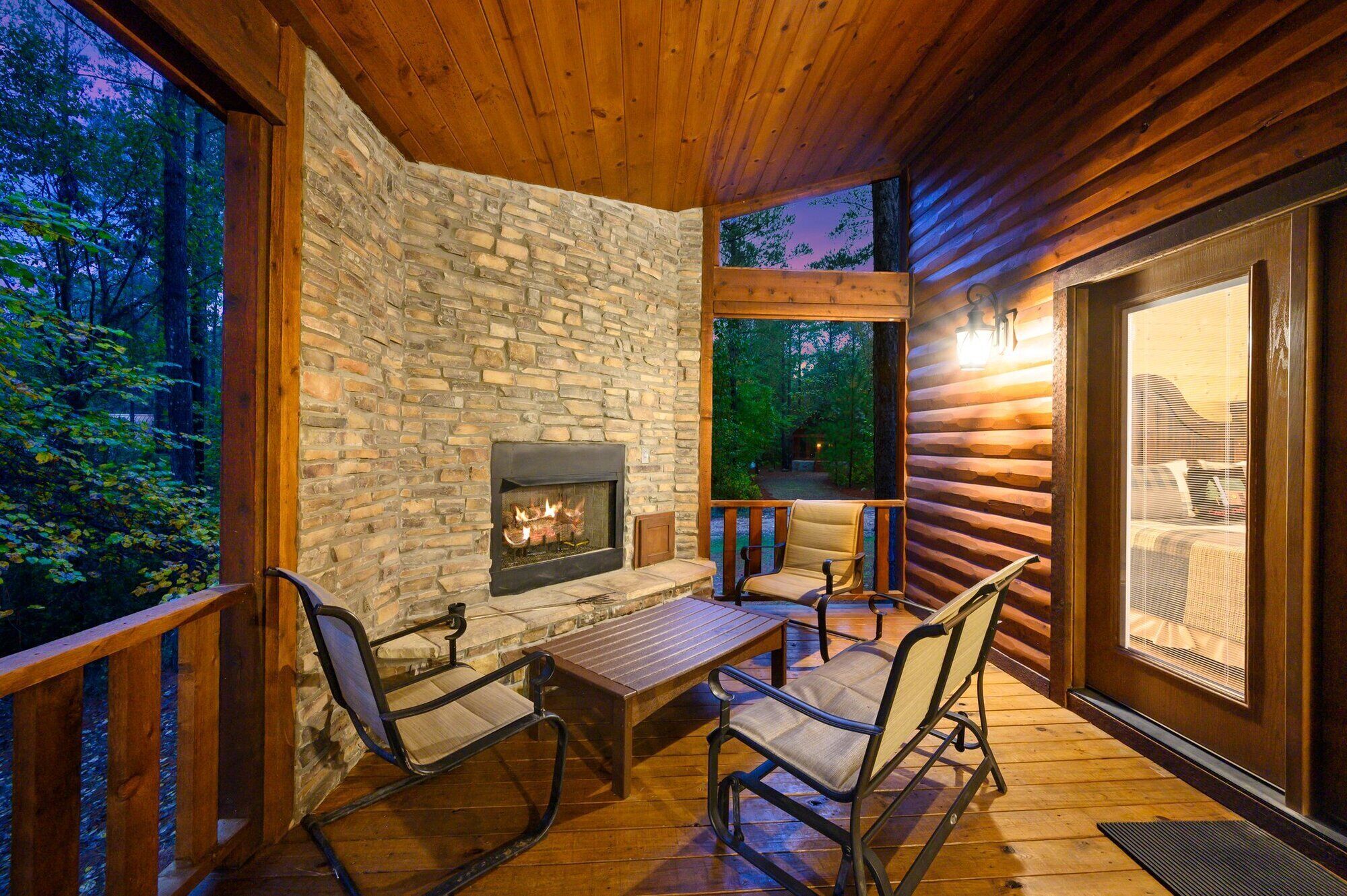 A wooden deck with a stone fireplace, seating, and a view of the forest.