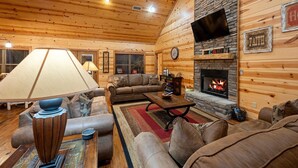 A cozy cabin interior with a stone fireplace, wooden walls, a leather sofa, and a television mounted above it.