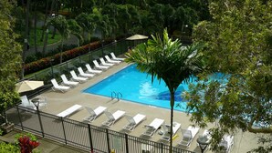 A pool area with sun loungers and umbrellas, surrounded by a fence and palm trees.