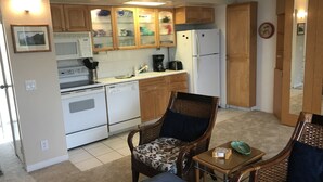 A compact kitchen with wooden cabinets, a microwave, a refrigerator, and a dishwasher. There are two wicker chairs with floral upholstery and a small table with a green bowl.