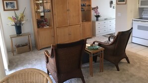 A living room with a wooden cabinet, wicker chairs, a small table, and a kitchen area with a microwave.