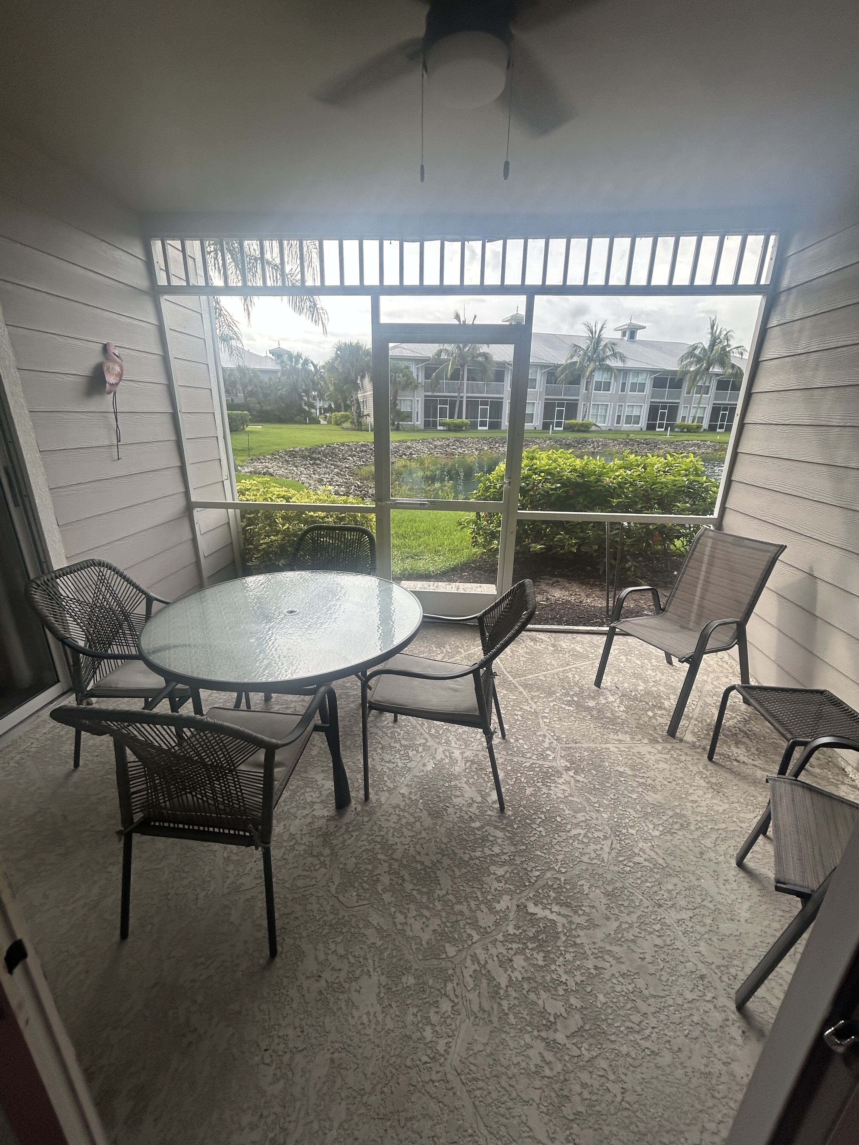 A covered patio area with a round glass table and four chairs, overlooking a landscaped garden and a building with multiple windows.