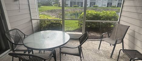 A covered patio area with a round glass table and four chairs, overlooking a landscaped garden and a building with multiple windows.