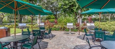 An outdoor patio area with green chairs and tables, surrounded by lush greenery and equipped with barbecue grills.